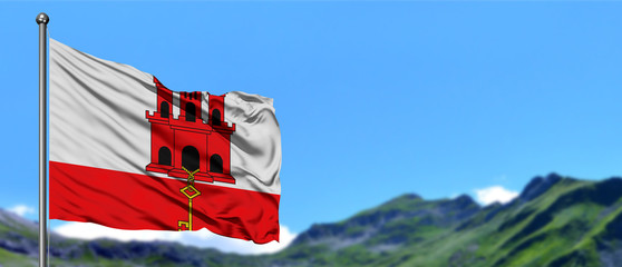 Gibraltar flag waving in the blue sky with green fields at mountain peak background. Nature theme.