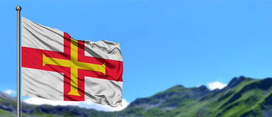 Guernsey flag waving in the blue sky with green fields at mountain peak background. Nature theme.