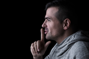 Studio portrait of a man looking sideways, with finger on lips. Isolated on black background. Horizontal. Copyspace.