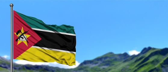 Mozambique flag waving in the blue sky with green fields at mountain peak background. Nature theme.