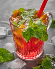 A light carbonated cherry drink with orange slices and pieces of ice, mint leaves and a cocktail tube to quench thirst on a hot day, pieces of ice are scattered alongside on the table.