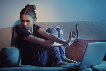 Frightened teenage girl with laptop on sofa in dark room. Danger of internet