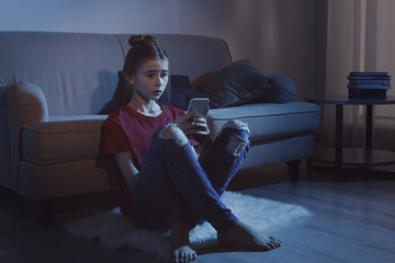 Upset teenage girl with smartphone in dark room. Danger of internet