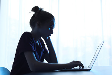 Upset teenage girl with laptop at table indoors. Danger of internet
