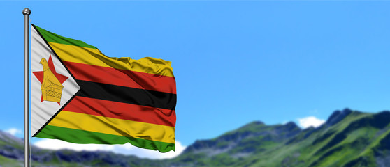 Zimbabwe flag waving in the blue sky with green fields at mountain peak background. Nature theme.