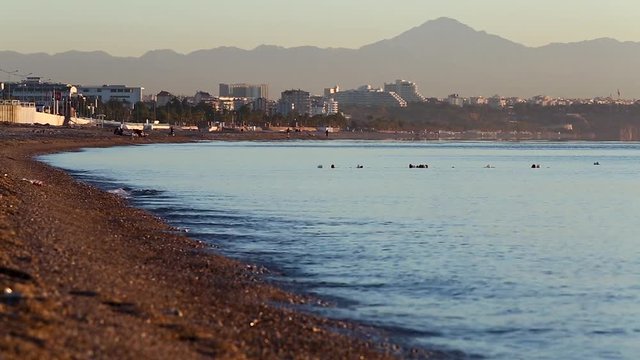 man running on the public beach with calm sea at the sunrise in Antalya, Turkey. small waves roll on the seashore with pebbles. beautiful morning sky over the sea and beach, part of Antalya city on