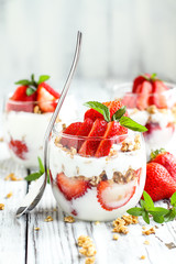 Healthy breakfast of strawberry parfaits made with fresh fruit, yogurt and granola over a rustic white table. Shallow depth of field with selective focus on glass jar in front. Blurred background.
