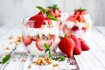 Healthy breakfast of strawberry parfaits made with fresh fruit, yogurt and granola over a rustic white table. Shallow depth of field with selective focus on glass jar in front. Blurred background. - obrazy, fototapety, plakaty