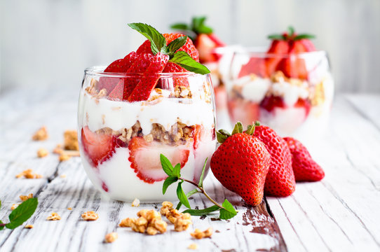 Healthy breakfast of strawberry parfaits made with fresh fruit, yogurt and granola over a rustic white table. Shallow depth of field with selective focus on glass jar in front. Blurred background.
