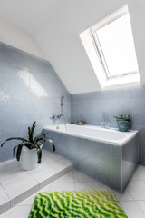 Bathroom corner in attic with bath, carpet, plants and roof window. Bath is equipped with modern stainless steel faucet. There can be seen a decorative accessories and walls are lined with blue tiles.