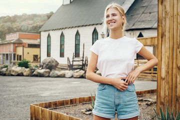 Young pretty girl is standing on a country village background. Woman is wearing a white empty t-shirt without logo which makes it being perfectly suitable for mock-up.