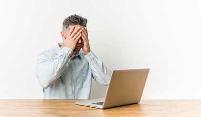 Young handsome man working with his laptop covers eyes with hands, smiles broadly waiting for a surprise.