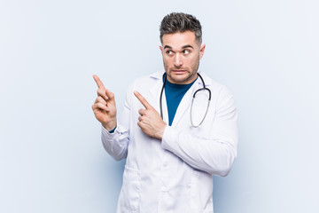 Young handsome doctor man shocked pointing with index fingers to a copy space.