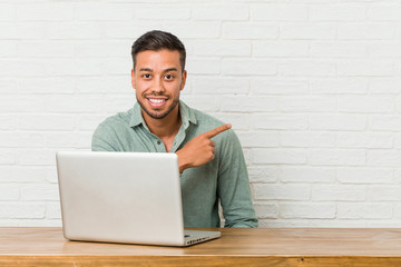 Young filipino man sitting working with his laptop smiling and pointing aside, showing something at blank space.