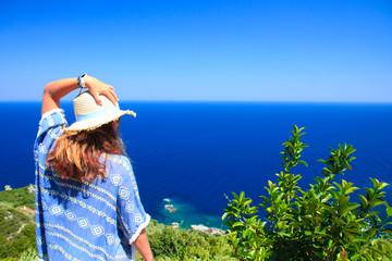 Female staring at the sea from a hill, while holding her hat with her left hand. Shot during daytime in summer
