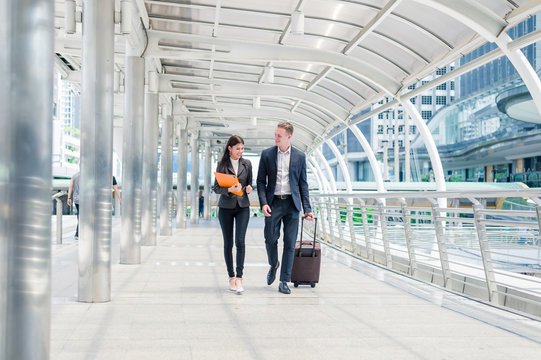 businessman with luggage and businesswoman with document file on hand walk together in the city, business travel concept