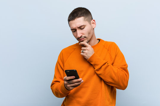 Young Caucasian Man Holding A Phone Looking Sideways With Doubtful And Skeptical Expression.