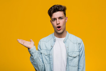 Young handsome student wearing a denim shirt impressed holding copy space on palm.