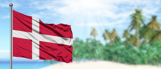 Waving Denmark flag in the sunny blue sky with summer beach background. Vacation theme, holiday concept.