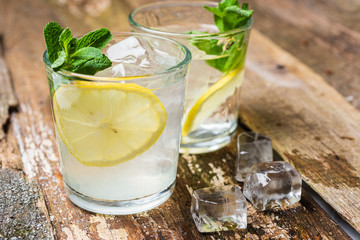 Two glasses of lemonade with with lemon slices, mint and ice on old wooden planks. Close up