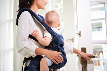 Young woman with little baby in sling opening front door and leaving house