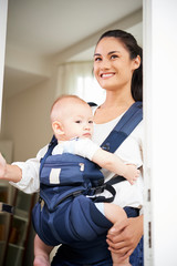 Happy young mother leaving house with adorable baby boy sitting in sling