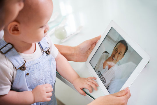 Mother sitting with baby on laps and video calling her husband via tablet computer