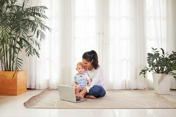 Young woman sitting floor with child on her laptop and answering e-mails on laptop