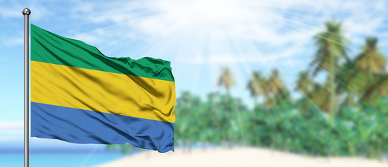 Waving Gabon flag in the sunny blue sky with summer beach background. Vacation theme, holiday...