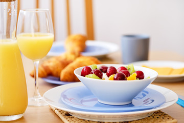 Close up fresh healthy nutritious breakfast jug glass with orange natural juice croissants on background, bowl plate with porridge or cereals garnished cherry, kiwi, orange slices on wicker place mat