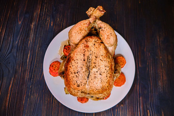 Whole grilled with carrot chicken on a plate. Festive dinner with meat bird. Fried juicy chicken on dark wooden background.