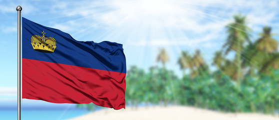 Waving Liechtenstein flag in the sunny blue sky with summer beach background. Vacation theme, holiday concept.