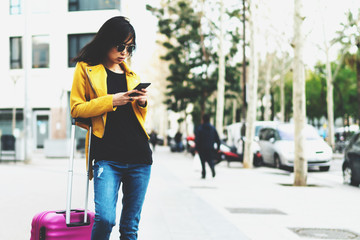 Asian hipster girl in trendy outfit booking hotel and flight tickets online by a mobile phone travel applications while standing with a suitcase on a street and waiting for a taxi to go to the airport