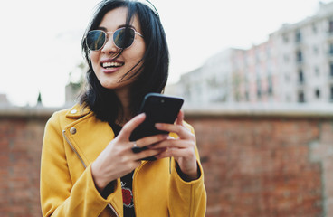 Smiling asian student girl dressed in casual wear looking away while posting photos to a social...