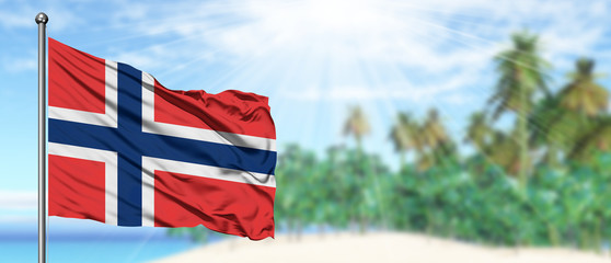 Waving Norway flag in the sunny blue sky with summer beach background. Vacation theme, holiday concept.