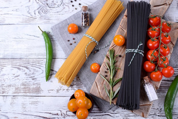 Different sorts of dry pasta on the wooden board surrounded by spices, cherry tomatoes and greenery