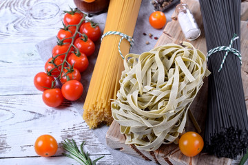 Different sorts of dry pasta on the wooden board surrounded by spices, cherry tomatoes and greenery