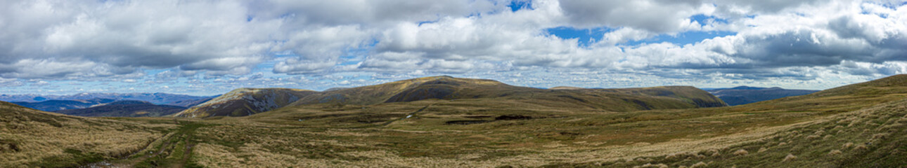 Fototapeta na wymiar A panorama view of a Scottish mountain summit plateau with heather and path trail under a majestic blue sky and huge white clouds