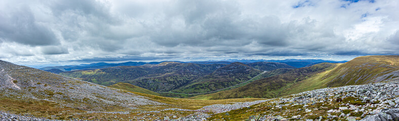 A panorama view of a Scottish mountain valley with a road and mountain range under a stormy huge white clouds sky