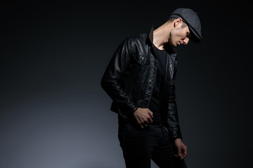 Fototapeta na wymiar Thoughtful man looking down upset and holding his hand on his leather jacket while wearing a hat and black jeans, posing on gray studio background