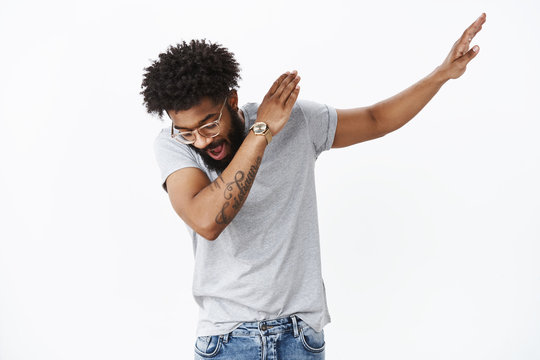 Swag in air. Portrait of handsome and stylish cool african american party guy with beard and tattoos sweep hand right and looking down as making dance move, shaking and having fun over gray wall