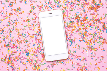 Mobile phone mock up and sweet multicolored sprinkles on pink pastel table in flat lay style. Fashion colour