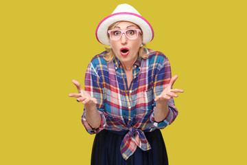 Portrait of surprised modern stylish mature woman in casual style with hat and eyeglasses standing, raised arms, looking at camera with shocked face. indoor studio shot isolated on yellow background.