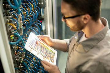 Close-up of concentrated man in shirt standing by mainframe and using digital tablet to check...