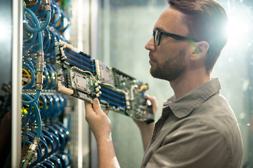 Serious skilled young bearded server installation specialist in glasses holding circuit board and...