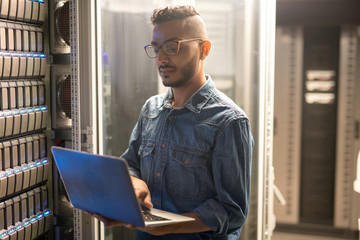 Serious concentrated young middle-eastern server specialist in glasses standing at network server and using portable computer while doing network monitoring