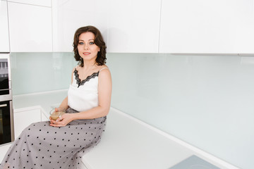 A young beautiful Caucasian woman sits at a table in the white modern kitchen, the housewife drinks coffee with milk.