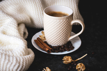 A cup of coffee is the key to a good mood. On a black, dark textural background, a composition of white, cream scarf and a white cup with espresso, coffee beans, cinnamon sticks and dry flowers