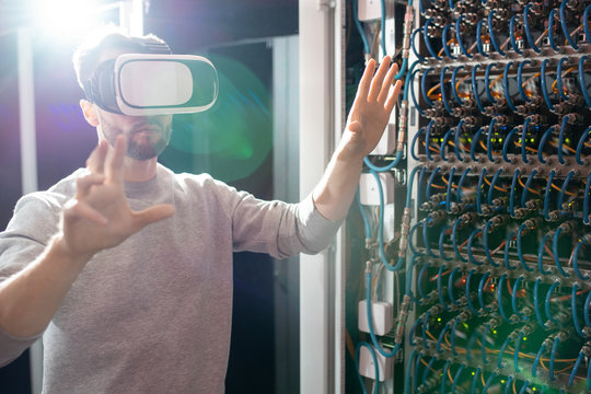 Serious concentrated young bearded engineer in VR goggles standing in database center and gesturing hands while managing server links wirelessly - Powered by Adobe