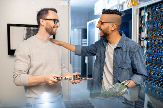 Positive excited young multi-ethnic IT engineers in casual outfits standing in server room and repairing motherboards together 